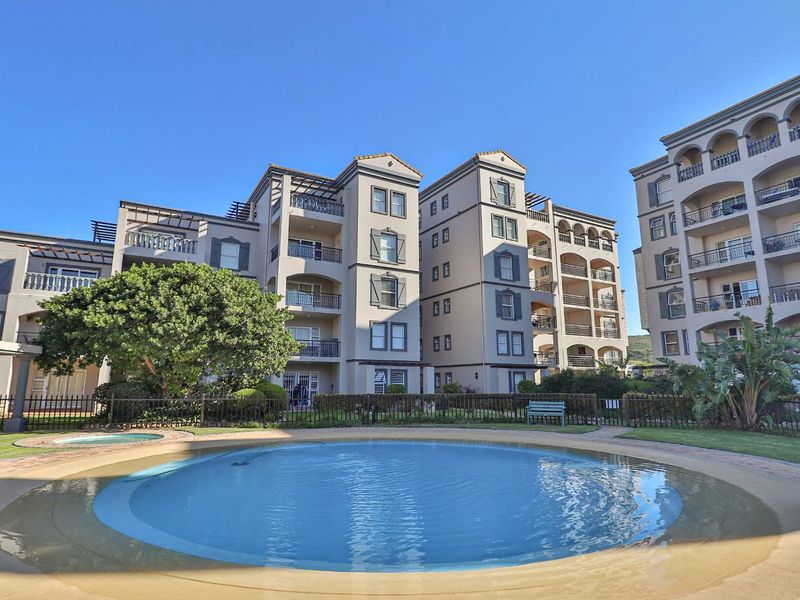 A well-maintained communal swimming pool area set within a multi-story apartment complex, featuring paved surrounds and landscaped garden borders.
