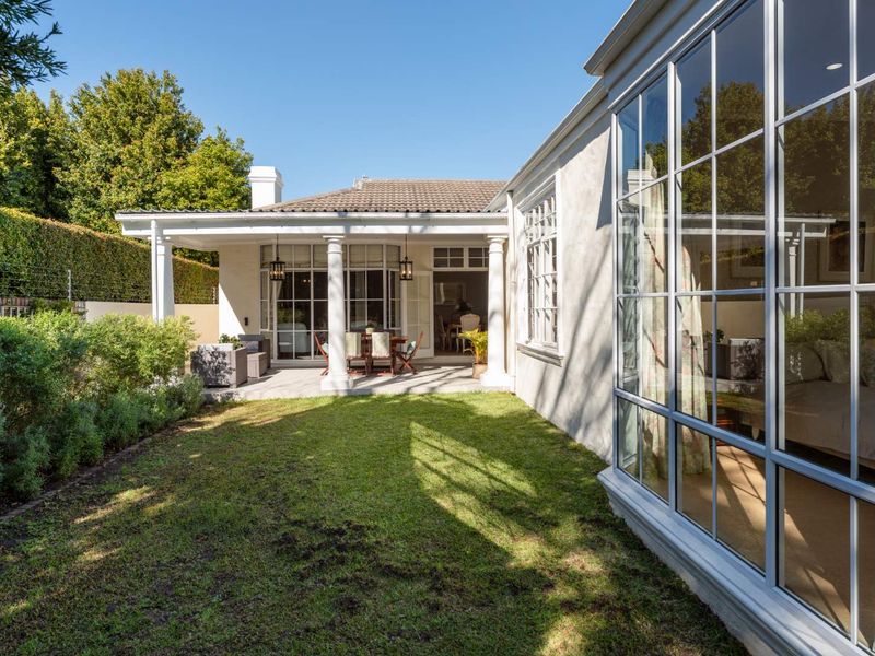 A well-maintained rear exterior view featuring a covered patio with classical columns, large grid-pattern windows, and a manicured garden.