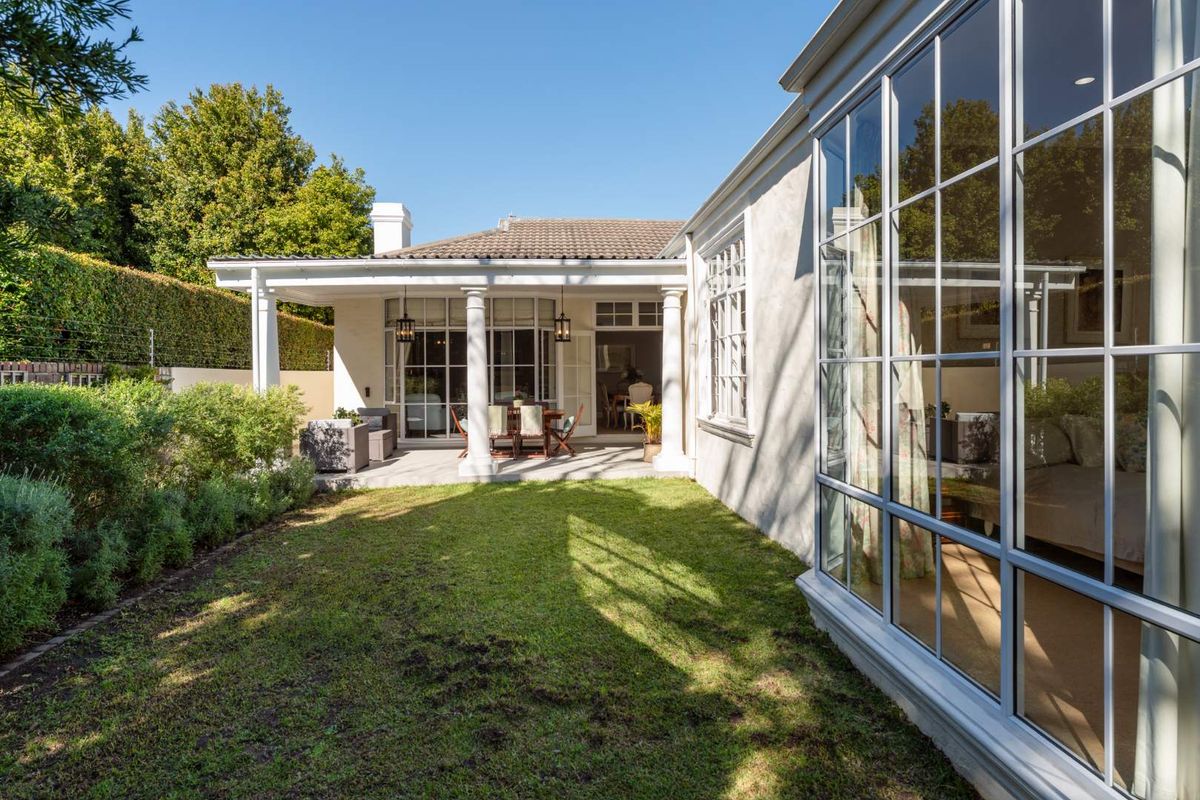 A well-maintained rear exterior view featuring a covered patio with classical columns, large grid-pattern windows, and a manicured garden.