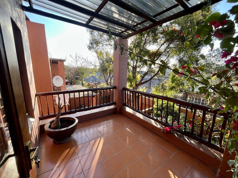 A covered balcony featuring tiled flooring, a wooden balustrade, and a corrugated roof, offering an outdoor space with views of the surrounding trees.