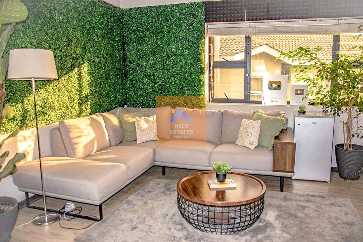 A cozy living room featuring laminate flooring, an artificial green wall feature, and large aluminium-framed windows allowing for natural light.