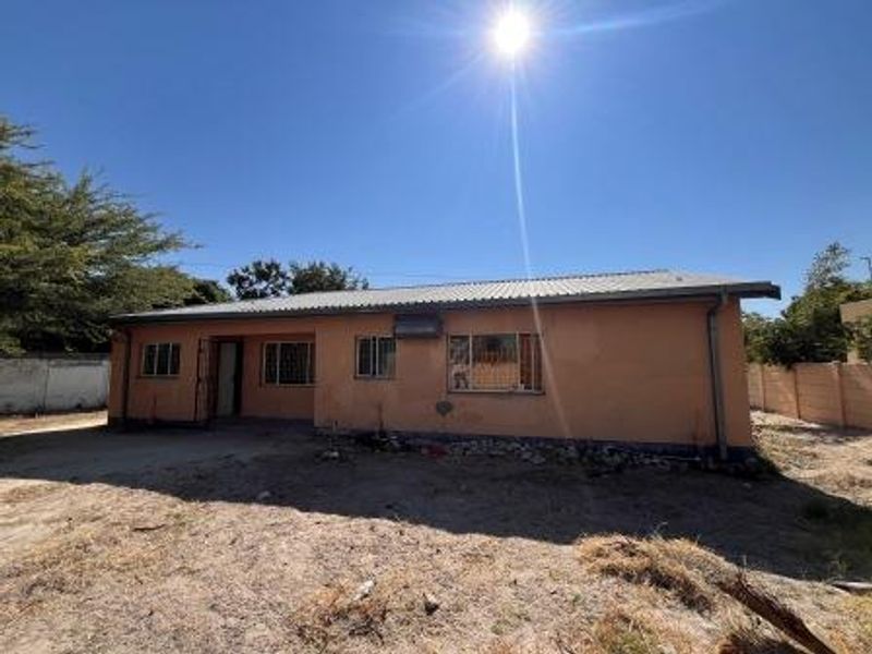 Rear view of a single-story house featuring plastered walls, a corrugated metal roof, and security bars on the windows, set on an unlandscaped plot.
