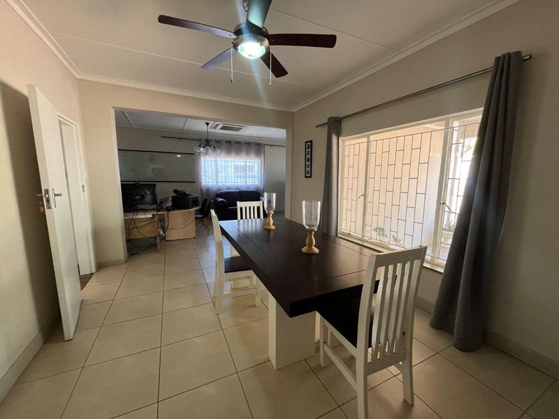 Open plan dining area featuring tiled flooring, a ceiling fan, and large windows with security bars, connecting to an adjacent living or office space.
