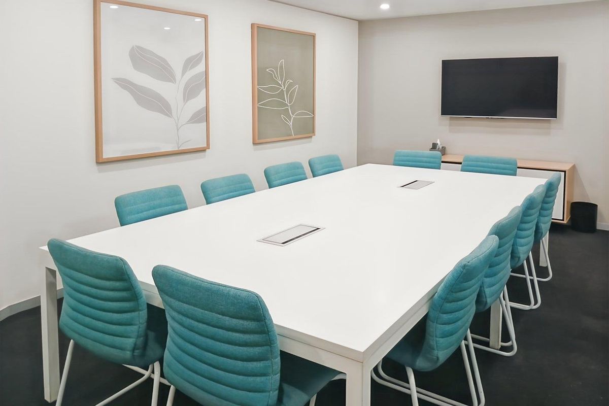A modern, well-lit office boardroom featuring a large central meeting table, recessed ceiling lighting, and a wall-mounted television for presentations.