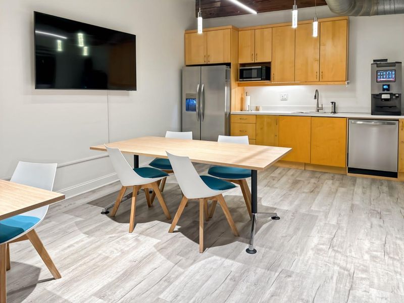 Modern open-plan office kitchenette featuring light wood cabinetry, stainless steel appliances, laminate flooring, and exposed industrial ceiling elements.