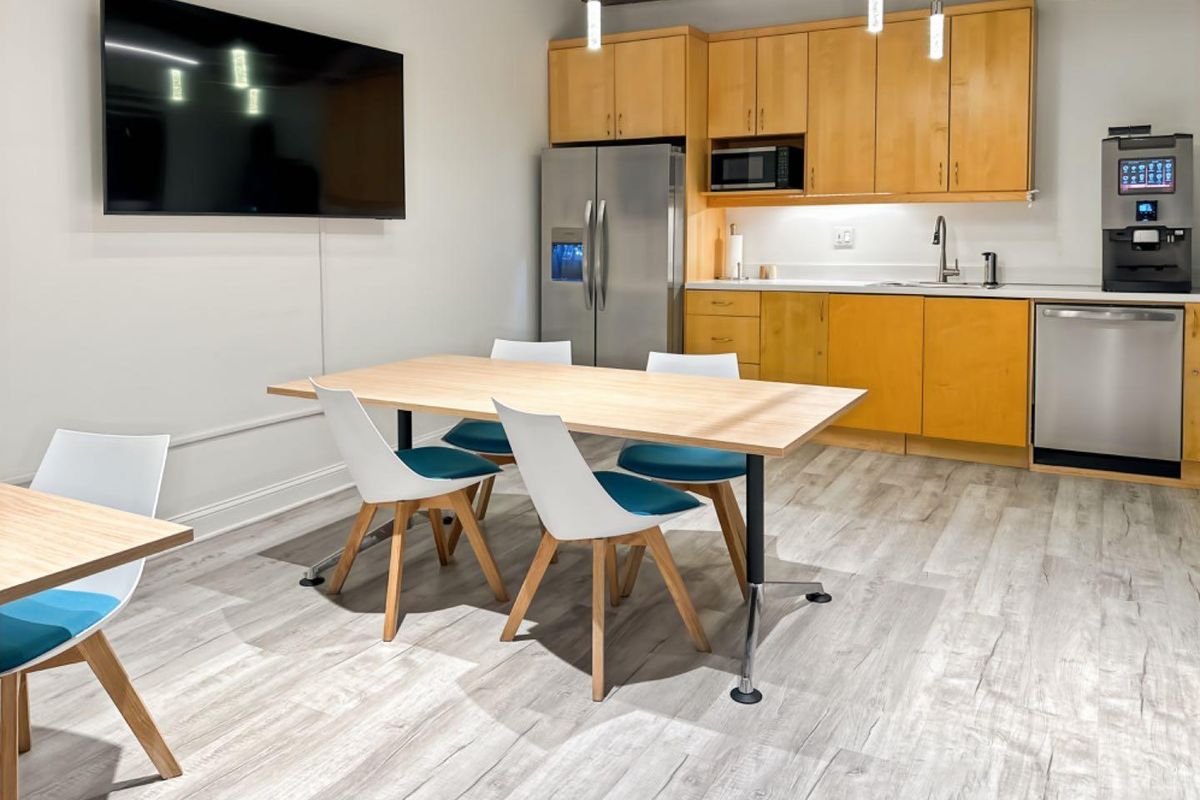 Modern open-plan office kitchenette featuring light wood cabinetry, stainless steel appliances, laminate flooring, and exposed industrial ceiling elements.
