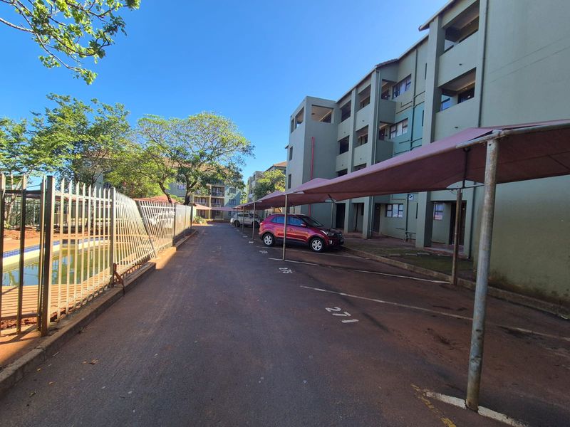 Exterior view of an apartment complex featuring covered parking bays, asphalt driveway, and communal swimming pool area with metal security fencing.