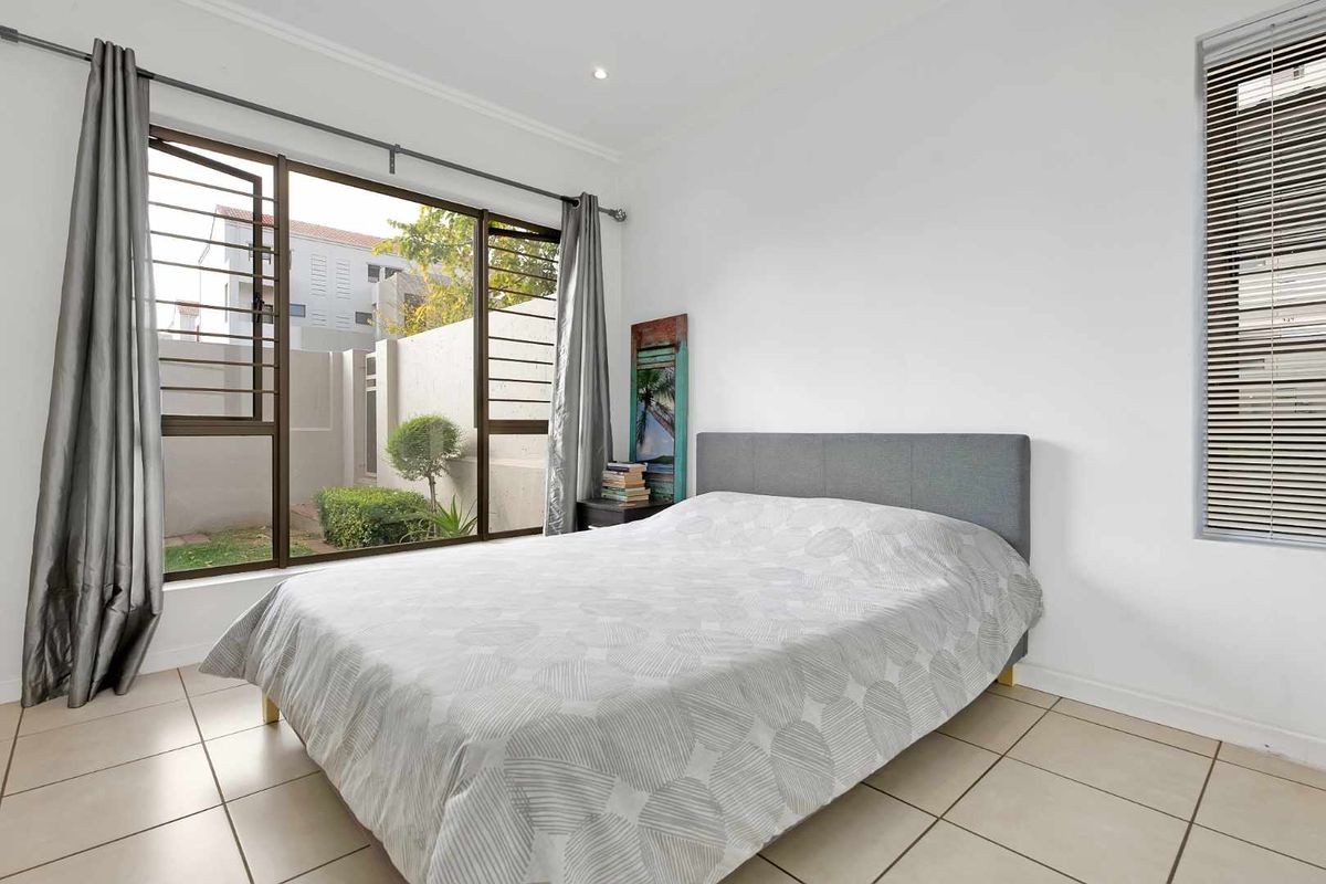 A bright bedroom featuring large windows with security bars, tiled flooring, and neutral wall finishes, offering a view of the exterior garden area.