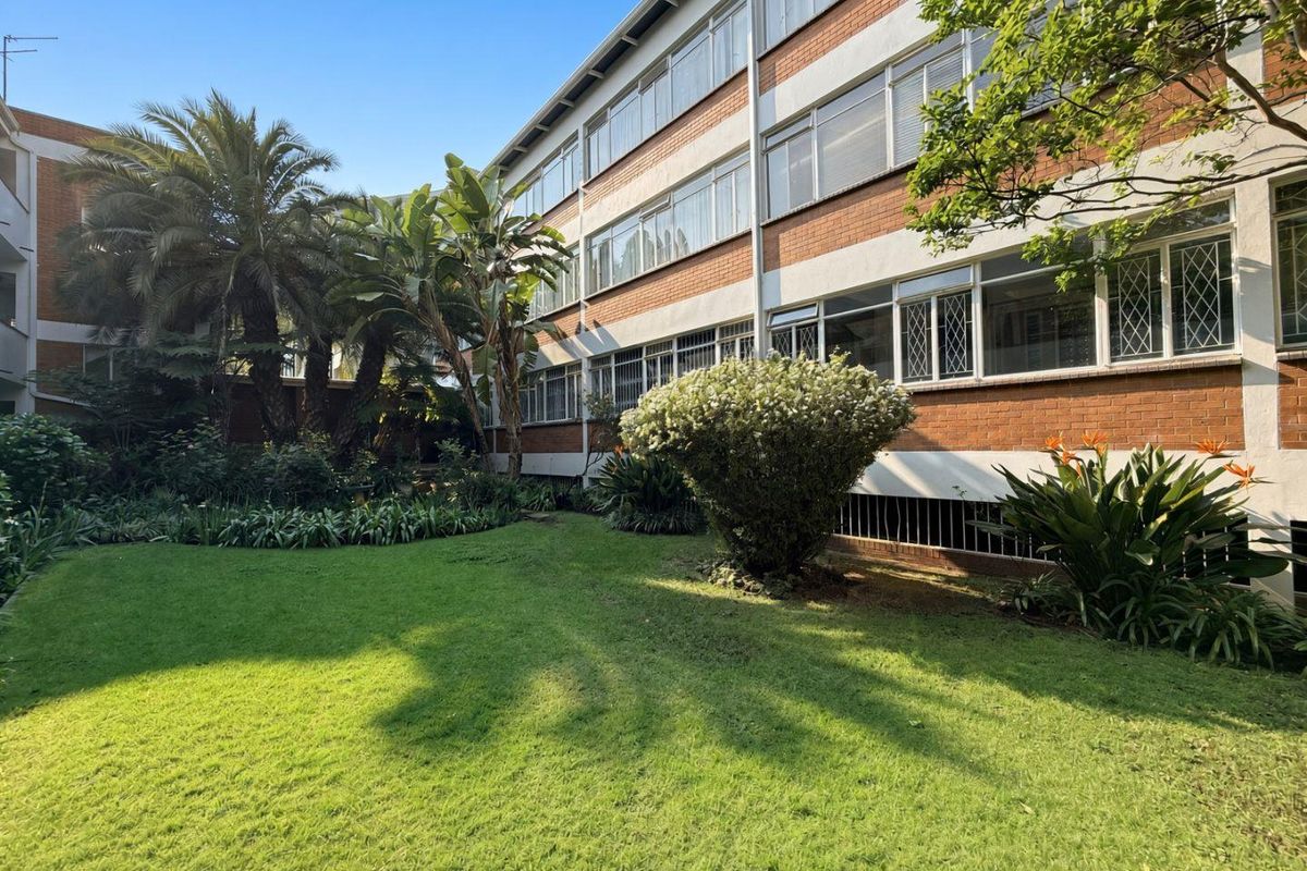 A well-maintained rear exterior view of the apartment building featuring a lush green garden, lawn, and classic brick facade with large windows.