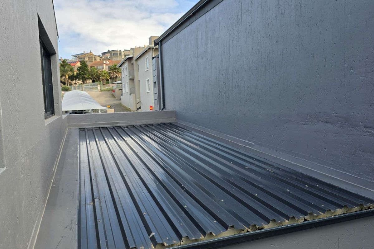 An exterior view of a corrugated metal roof section between building walls, showing modern grey exterior finishes and structural detailing.