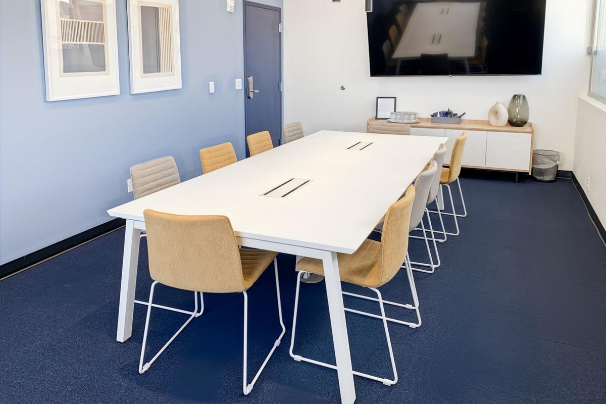 A modern, well-maintained office meeting room featuring blue accent walls, carpeted flooring, built-in cabinetry, and a wall-mounted television for presentations.