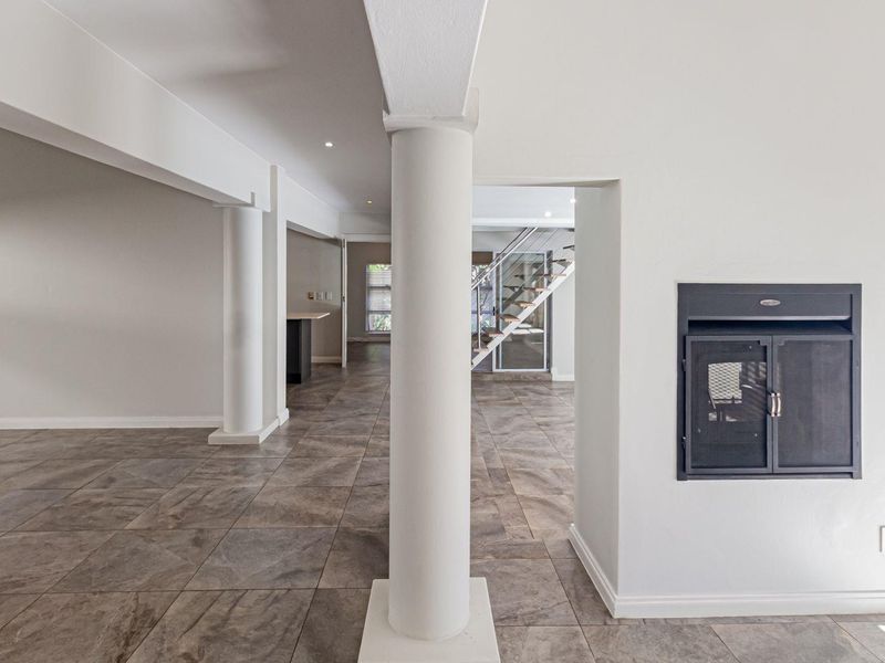Spacious open-plan living area featuring large format floor tiles, structural columns, a built-in fireplace, and a modern staircase with a glass balustrade.