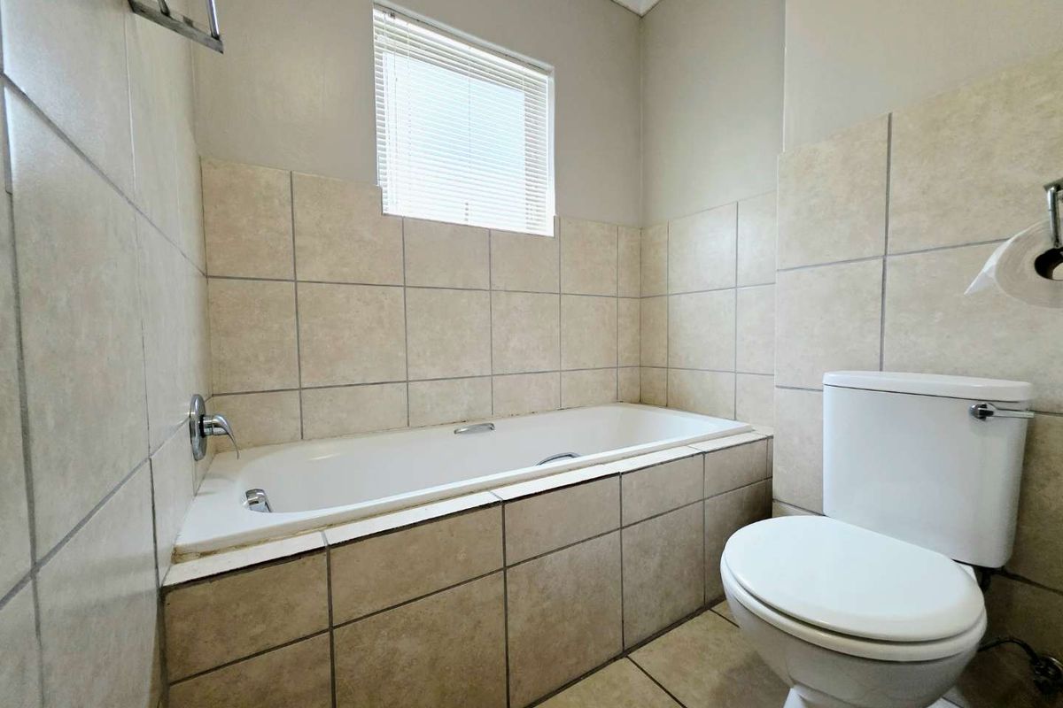 A functional bathroom featuring a built-in bathtub with tiled surround, a standard toilet, and a window with fitted blinds.