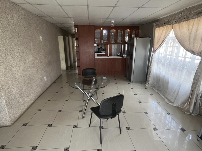 Open plan living and dining area featuring tiled flooring, drop ceiling, and built-in kitchen cabinetry with a security gate on the window.