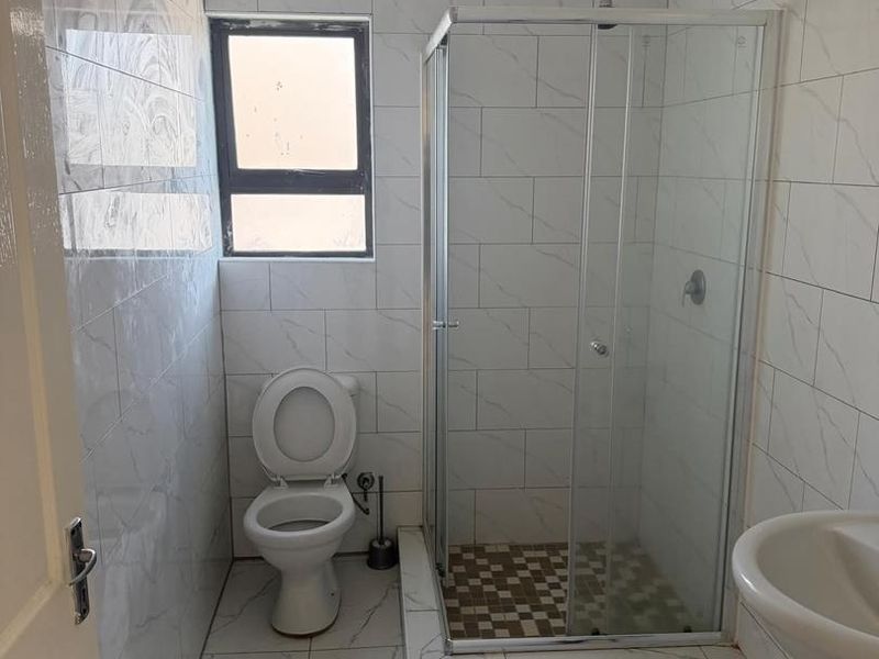 A clean, functional bathroom featuring a glass shower enclosure, pedestal basin, and toilet with modern white marble-look wall and floor tiling.