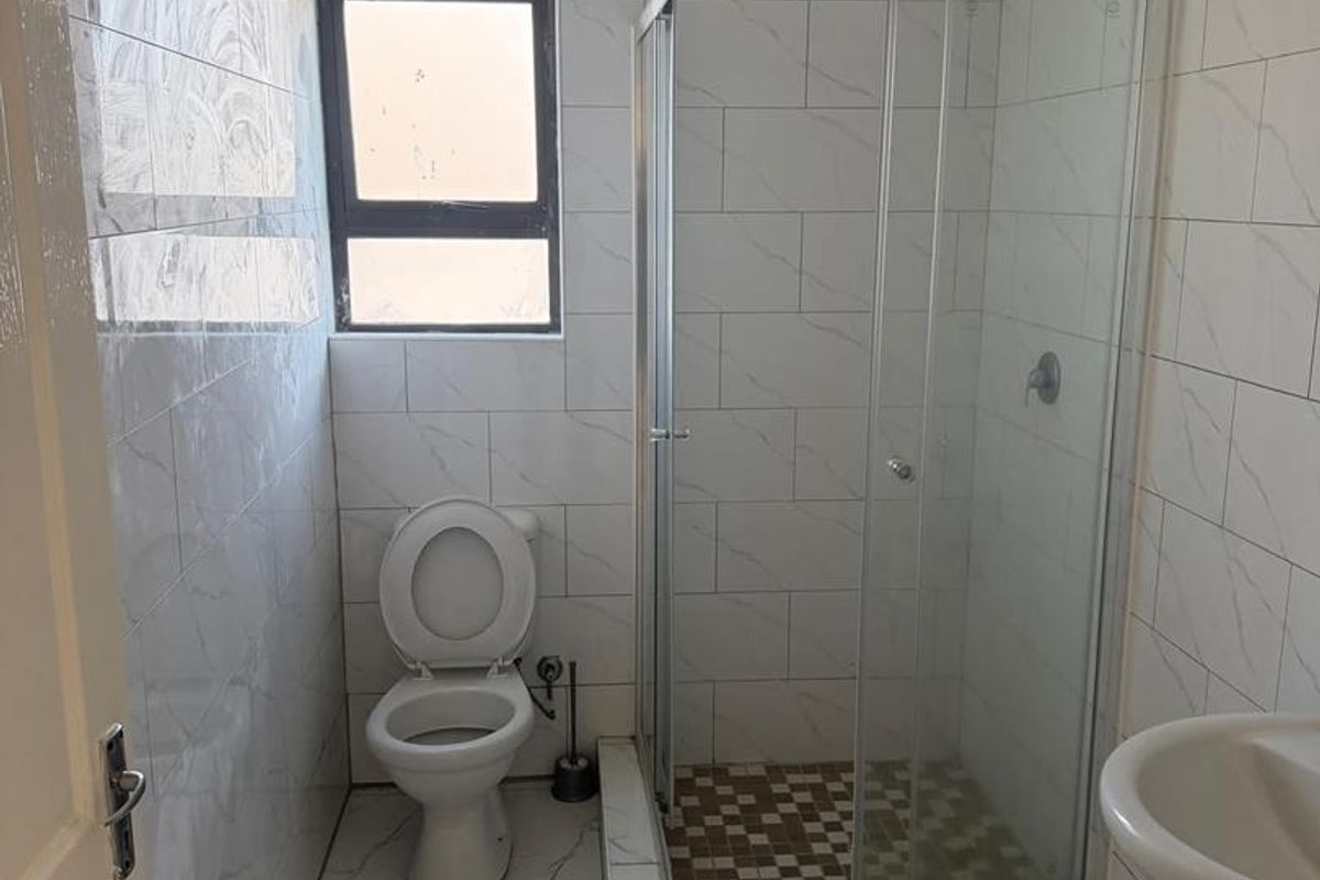 A clean, functional bathroom featuring a glass shower enclosure, pedestal basin, and toilet with modern white marble-look wall and floor tiling.