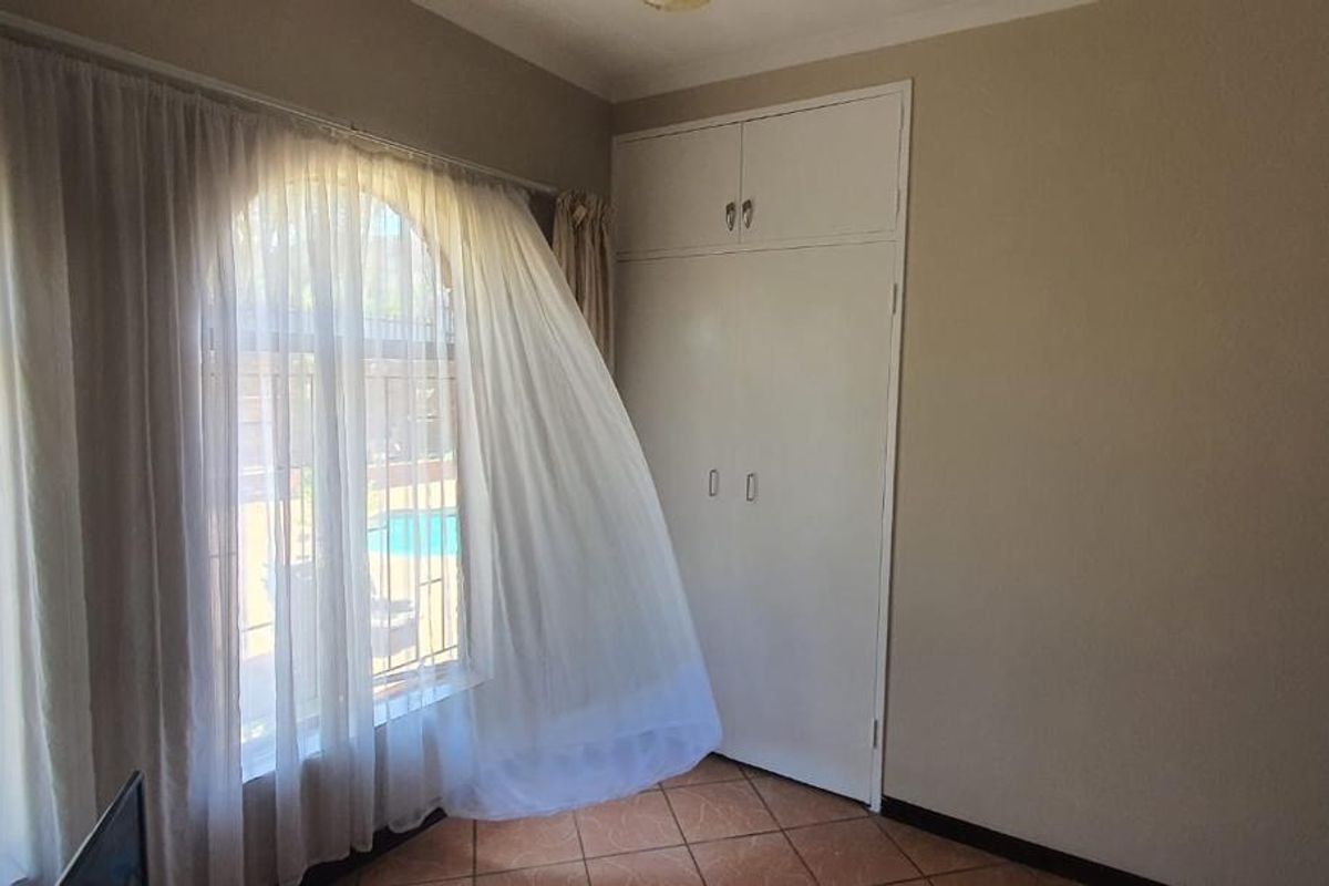 A bedroom featuring built-in cupboards, tiled flooring, and large arched windows with security bars overlooking the garden.