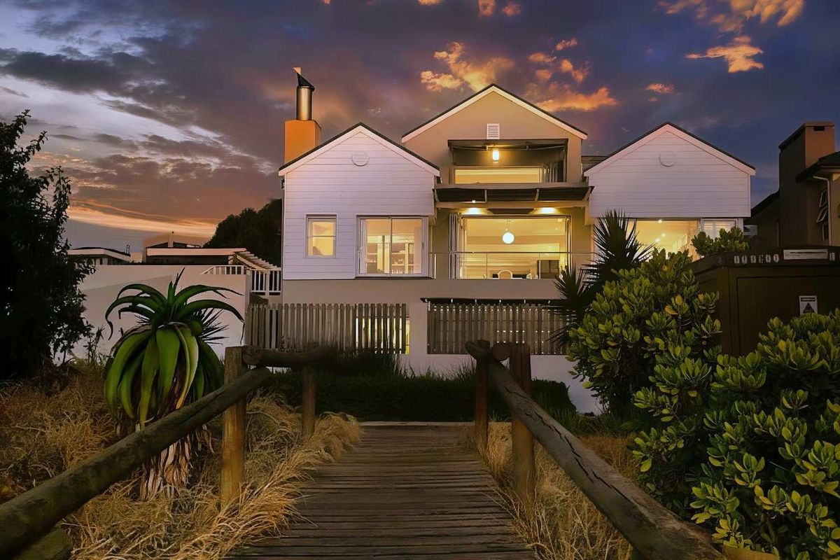 A stunning rear exterior view of a multi-level residence featuring a wooden boardwalk approach, balcony, and modern architectural design at sunset.