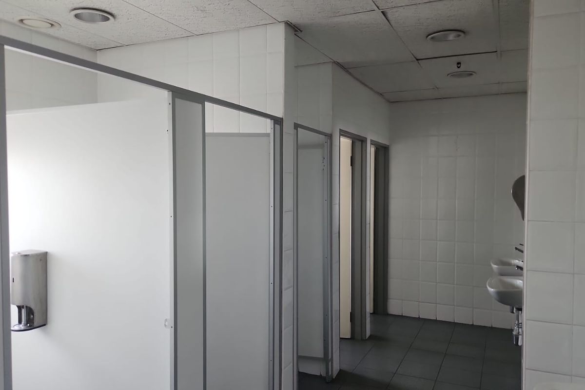 Functional industrial restroom facility featuring multiple partitioned cubicles, wall-mounted basins, and tiled surfaces throughout.