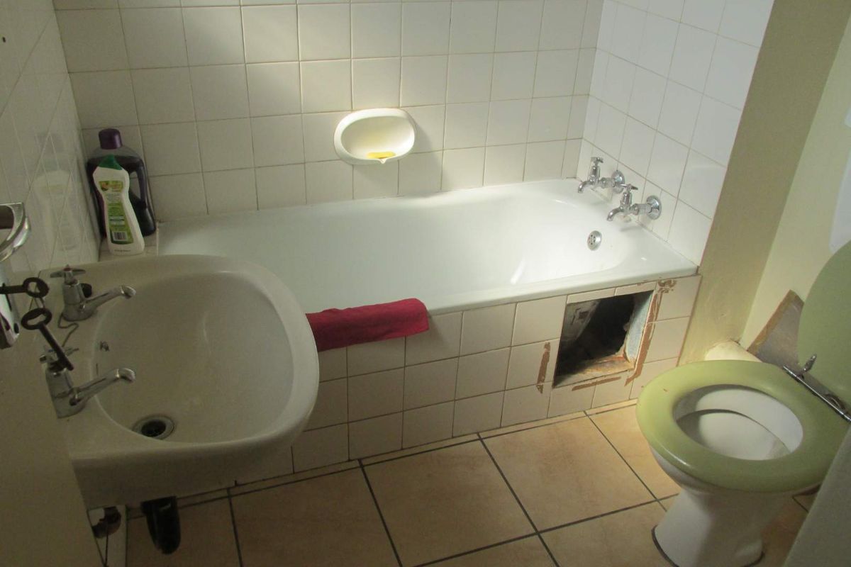 A dated bathroom featuring a standard bathtub, pedestal sink, and toilet, currently requiring maintenance and repair to the bath panel and tiling.
