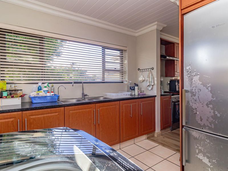 A functional kitchen featuring wooden cabinetry, granite countertops, a double sink, and tiled flooring, with a view into an adjacent scullery area.