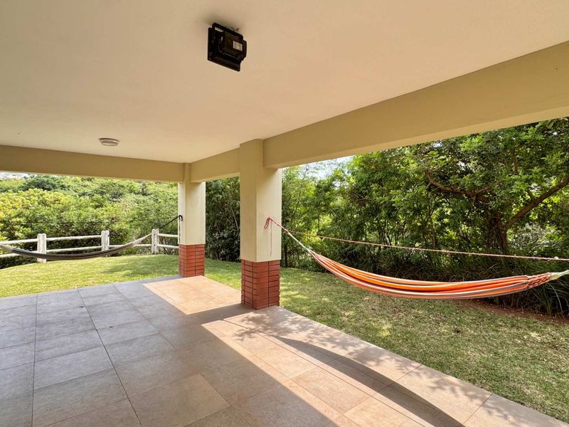 A spacious covered patio featuring tiled flooring and brick-based pillars, overlooking a lush green garden and natural landscape.