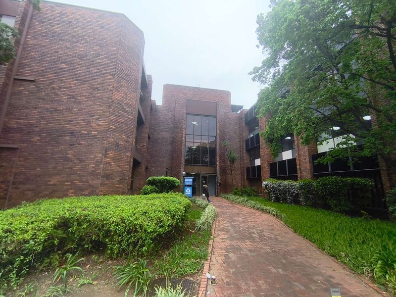 Front view of a multi-story brick office building featuring a paved walkway leading to a glass entrance, surrounded by established garden landscaping.