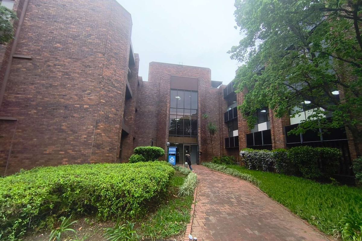 Front view of a multi-story brick office building featuring a paved walkway leading to a glass entrance, surrounded by established garden landscaping.