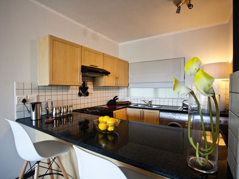 A functional kitchen featuring light wood cabinetry, dark granite countertops, a tiled backsplash, and integrated appliances including a hob and extractor fan.