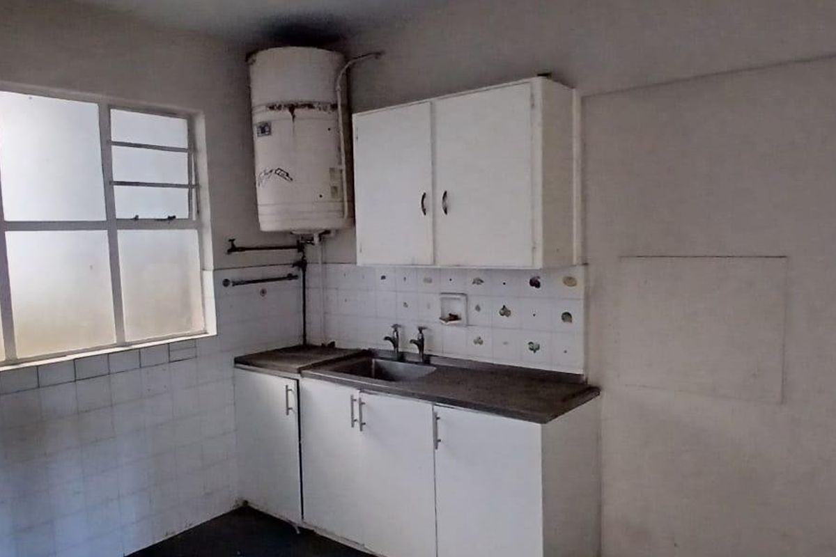 A basic, empty kitchen area featuring built-in cabinetry, a stainless steel sink, tiled splashbacks, and an exposed electric geyser unit.