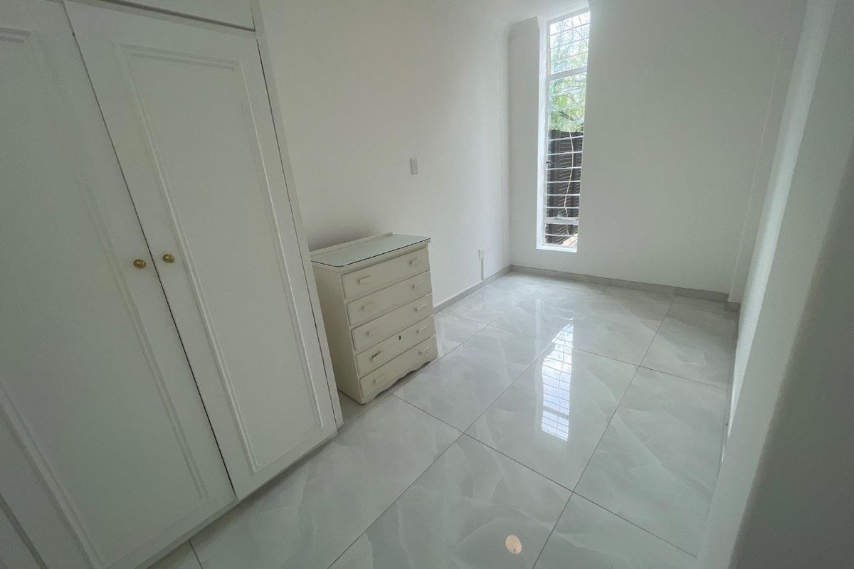 A bright bedroom featuring polished tiled flooring, a built-in wardrobe, and a tall window with security bars.