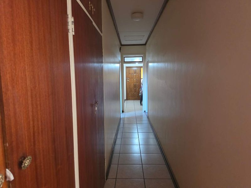 A long residential hallway featuring tiled flooring and built-in wooden cupboards along one side, leading to a doorway at the end.