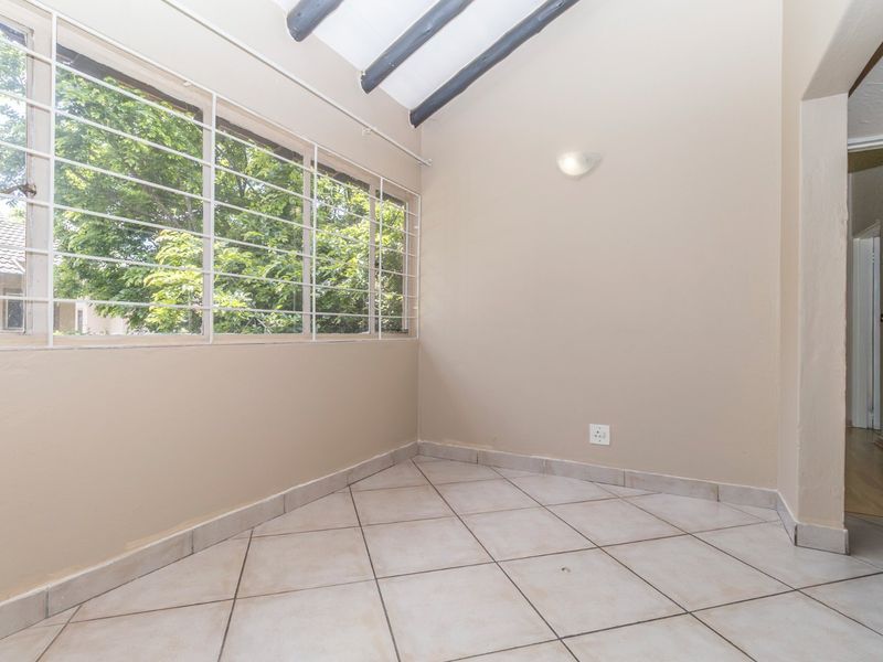 A sunlit, enclosed patio area featuring tiled flooring, exposed ceiling beams, and large windows with security bars, offering a versatile space for relaxation.
