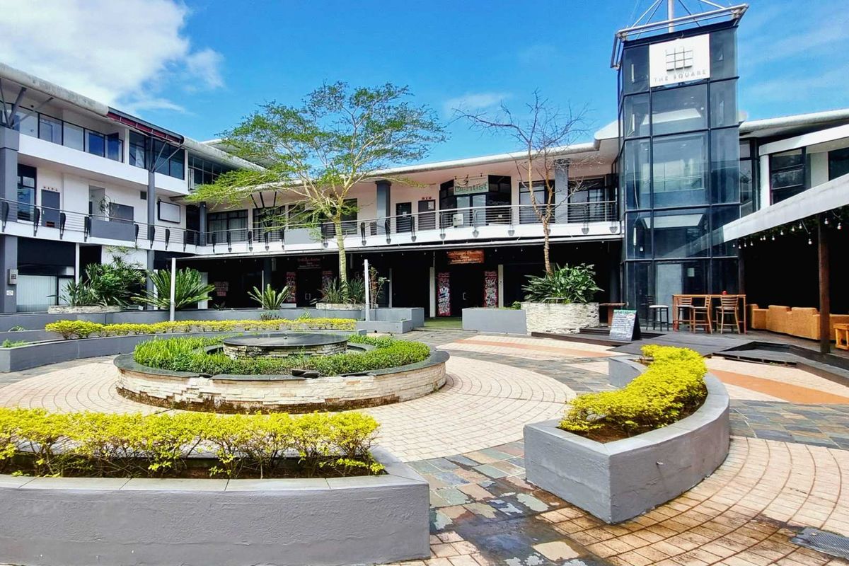 A central courtyard area within a mixed-use commercial complex, featuring paved walkways, landscaped garden beds, a central water feature, and a glass elevator shaft.