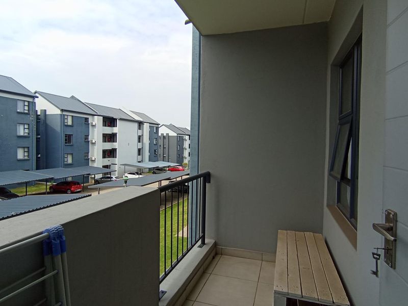 A clean, covered balcony featuring tiled flooring and a metal balustrade, overlooking the apartment complex parking area.