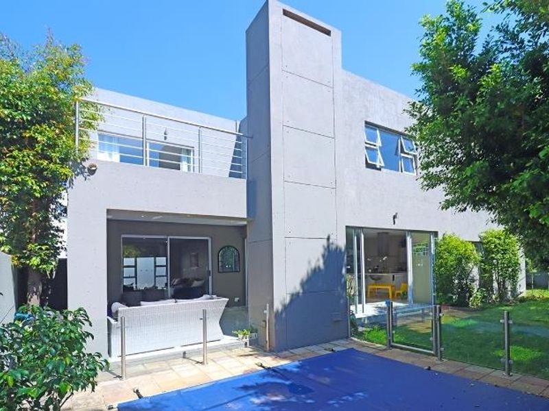 Rear view of a modern double-story cluster home featuring a swimming pool, paved patio area, and an upper-level balcony with stainless steel railings.