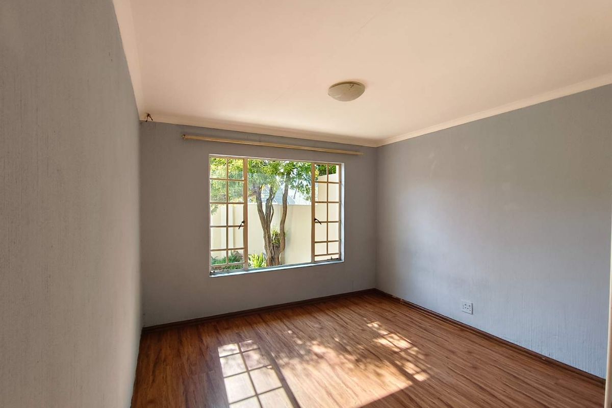 A bright, empty bedroom featuring laminate wood flooring, neutral grey walls, and a large steel-framed window overlooking the garden.