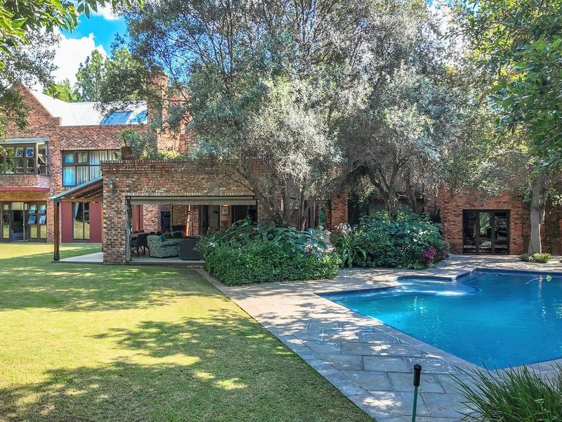 A spacious rear garden featuring a swimming pool, stone patio area, and a multi-level brick house with mature trees.