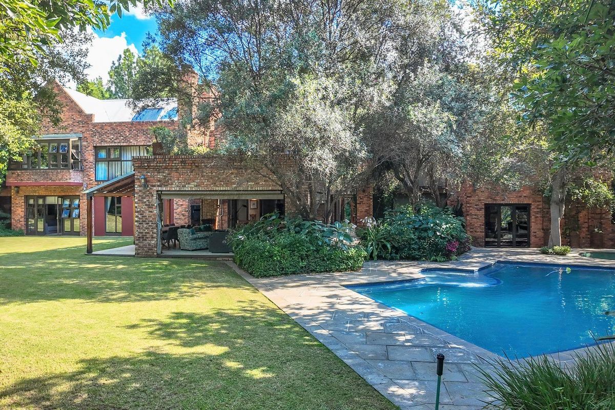 A spacious rear garden featuring a swimming pool, stone patio area, and a multi-level brick house with mature trees.