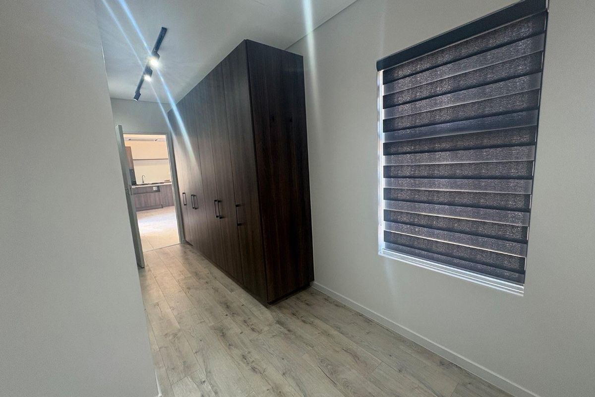 A modern, empty bedroom featuring dark wood built-in cupboards, light laminate flooring, and contemporary track lighting with roller blinds on the window.