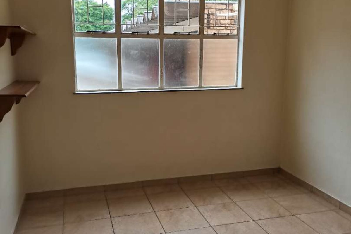 A simple, empty bedroom featuring tiled flooring, a large multi-pane window with security bars, and built-in wall shelving.