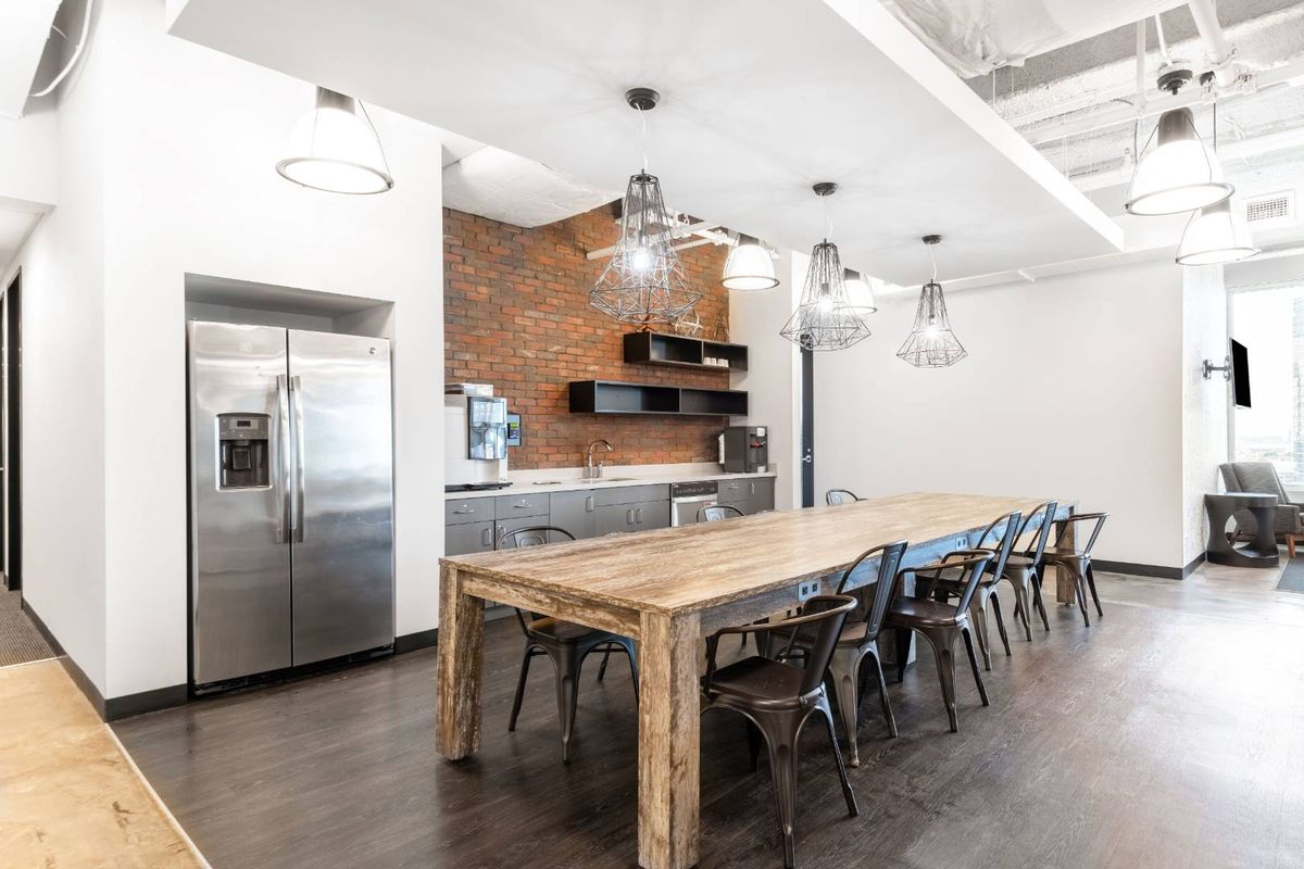 Modern office breakroom featuring a kitchenette with exposed brick backsplash, stainless steel appliances, and a large communal dining table.