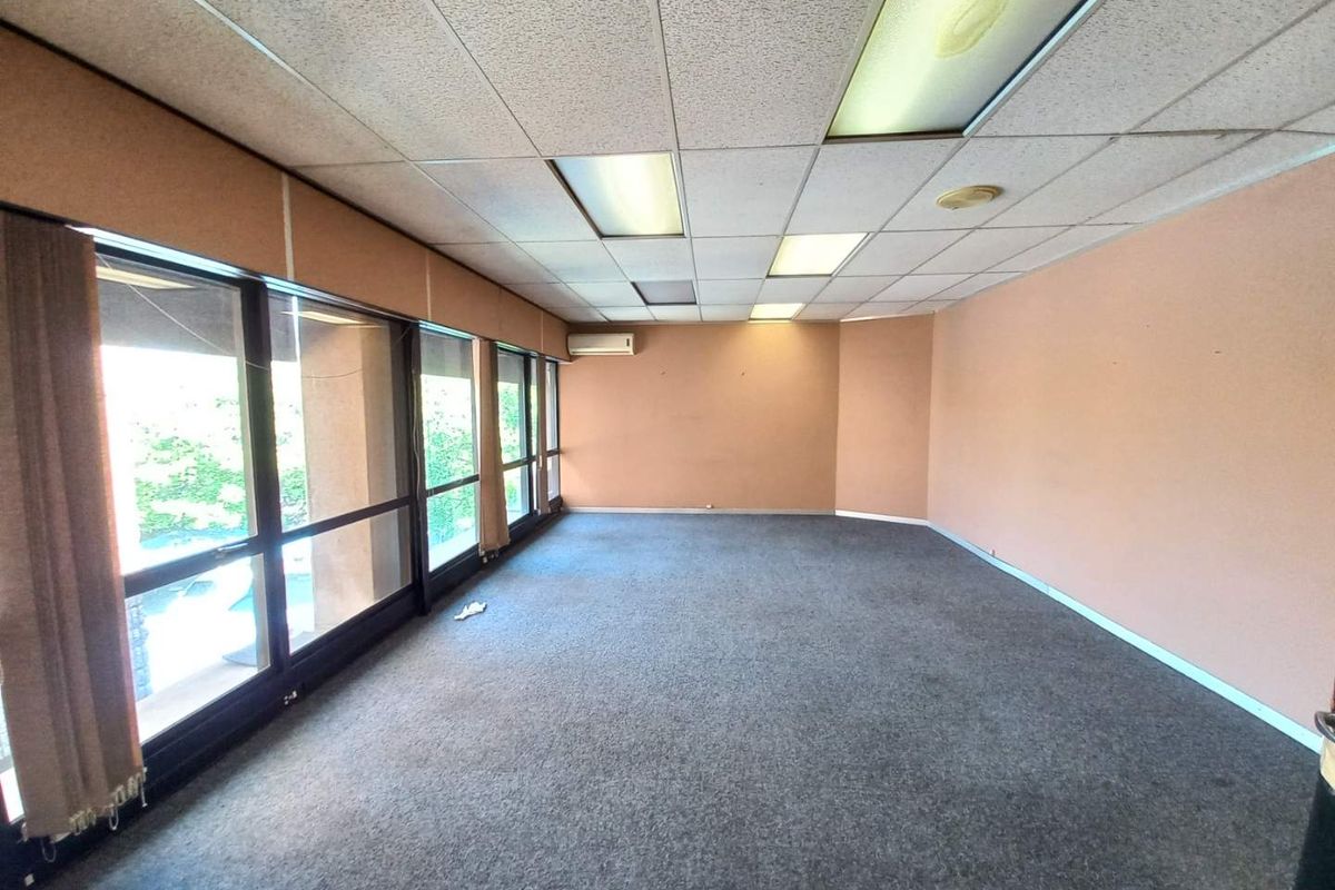 An empty office space featuring large windows, carpeted flooring, a drop ceiling with integrated lighting, and a wall-mounted air conditioning unit.