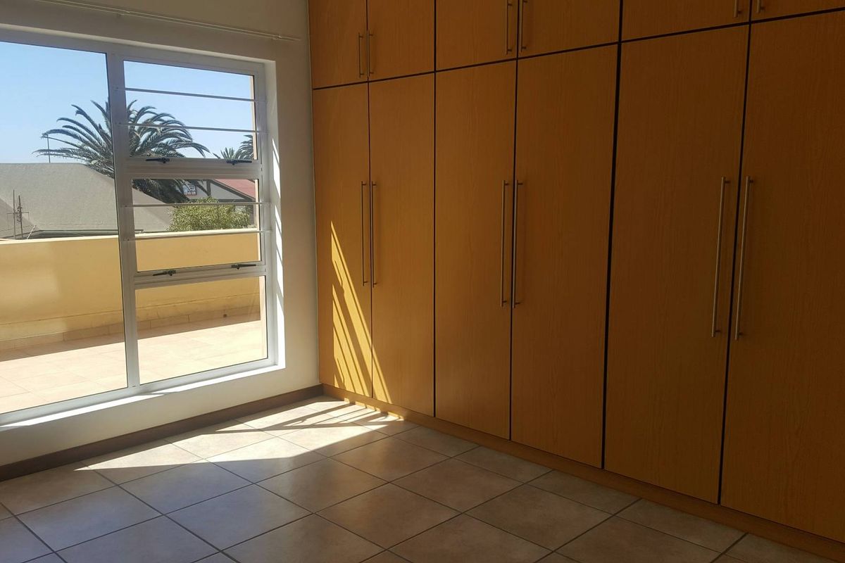 A bright, empty bedroom featuring extensive floor-to-ceiling built-in wooden cupboards, tiled flooring, and large windows with access to an outdoor balcony area.