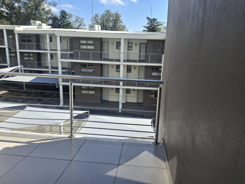 A clean, tiled balcony featuring a modern stainless steel balustrade, overlooking the apartment complex courtyard and covered parking area.