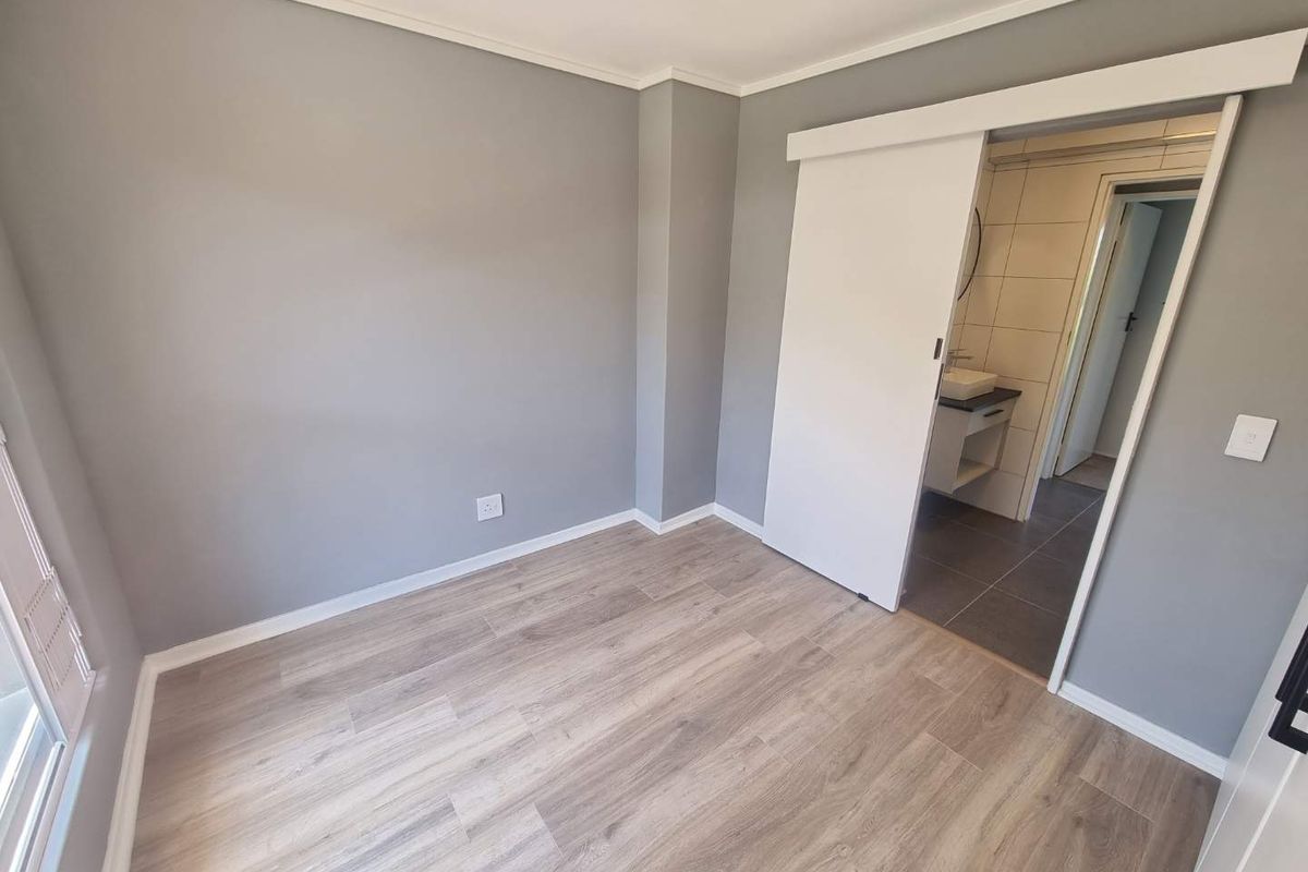 A bright, modern bedroom featuring light wood-look laminate flooring, neutral grey walls, and a sliding barn door leading to an en-suite bathroom.
