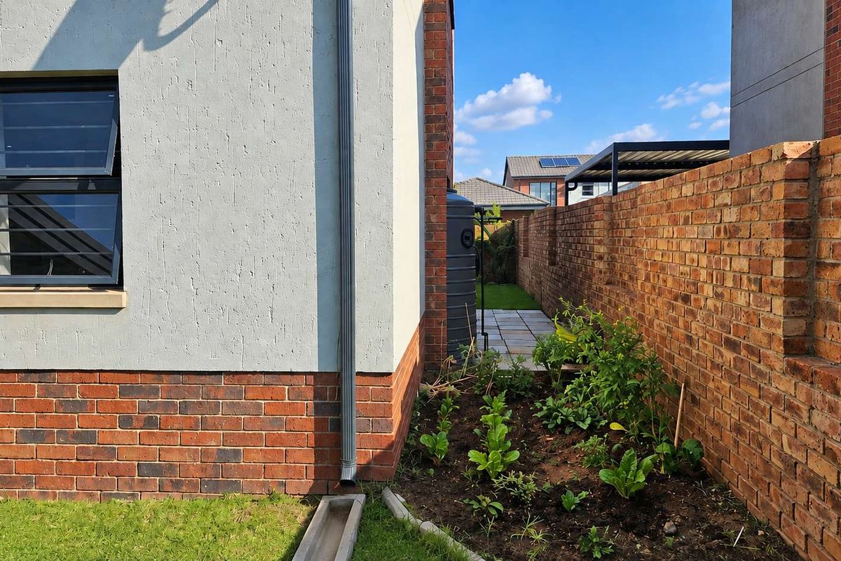 A side view of the property showing a brick boundary wall, paved walkway, rainwater downpipe, and a water storage tank in the garden area.