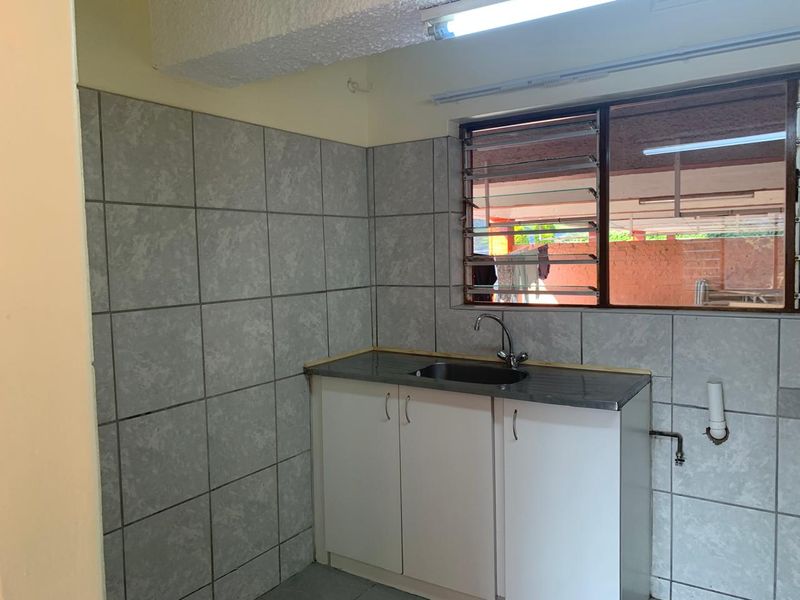 A compact kitchen area featuring a stainless steel sink, built-in white cabinetry, and grey tiled splashbacks with a louvered window.