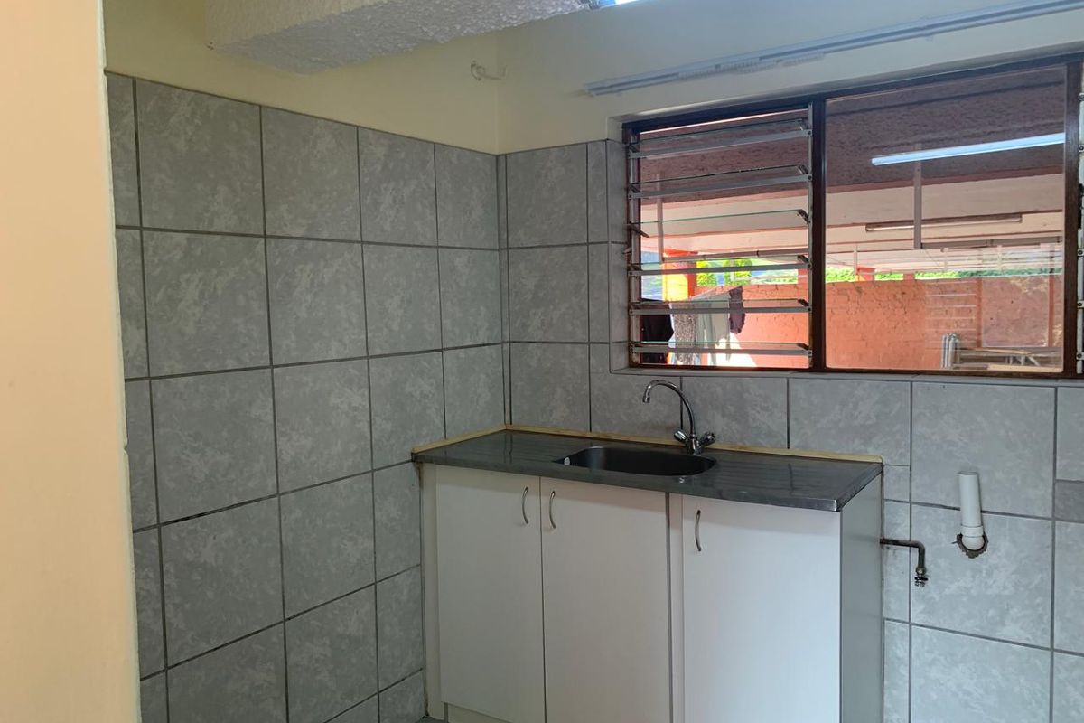 A compact kitchen area featuring a stainless steel sink, built-in white cabinetry, and grey tiled splashbacks with a louvered window.