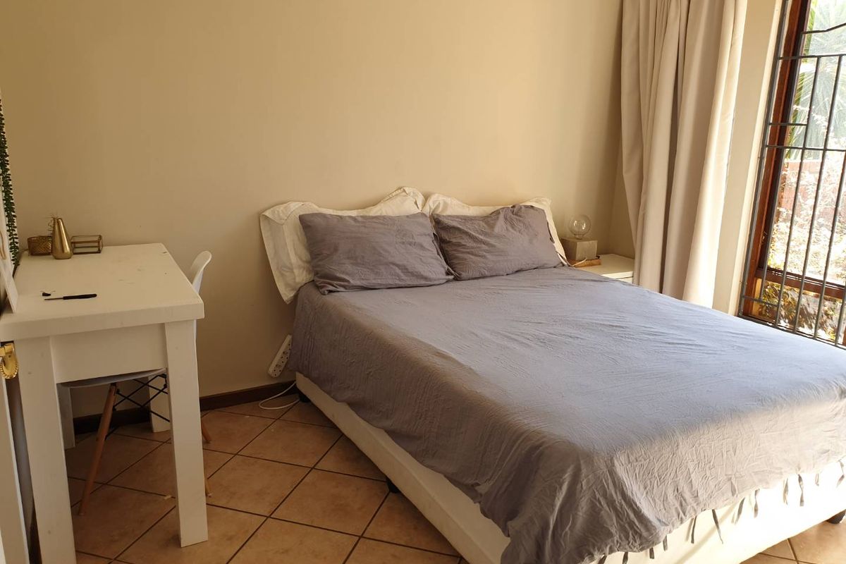 A functional bedroom featuring tiled flooring, neutral wall tones, and a large window with security bars, providing natural light and a view of the exterior.